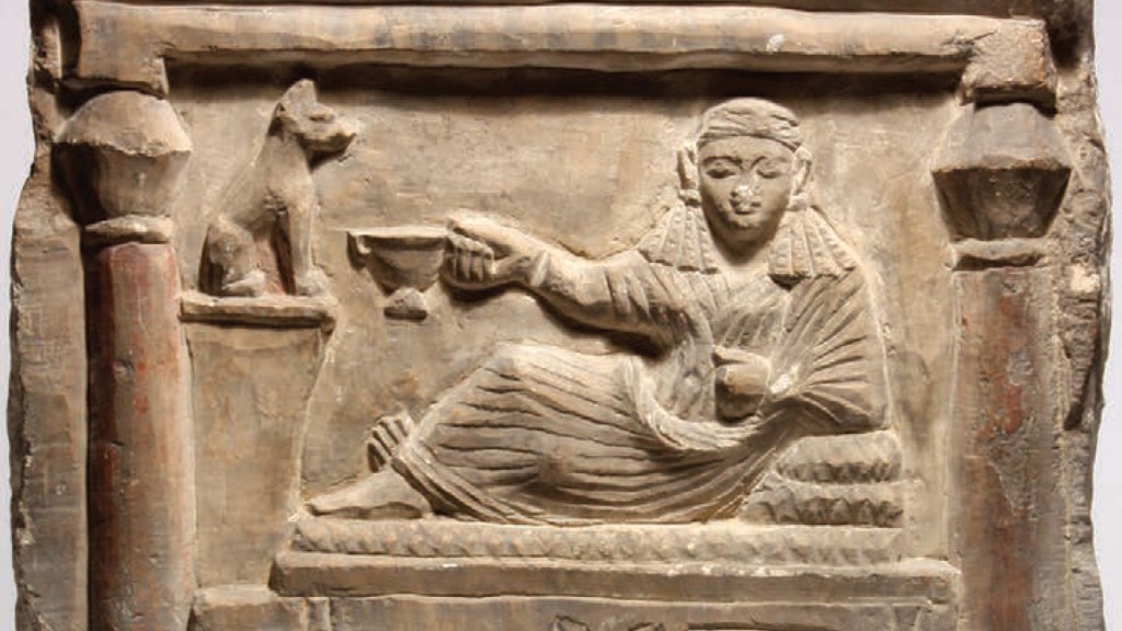 The stele shows a woman carved in stone in Egyptian aesthetic style. She wears a headdress. She faces forward but her legs are off to the side as if she is reclining. She holds a drinking vessel towards a cat who is sitting above her and to her left. She holds a musical instrument.